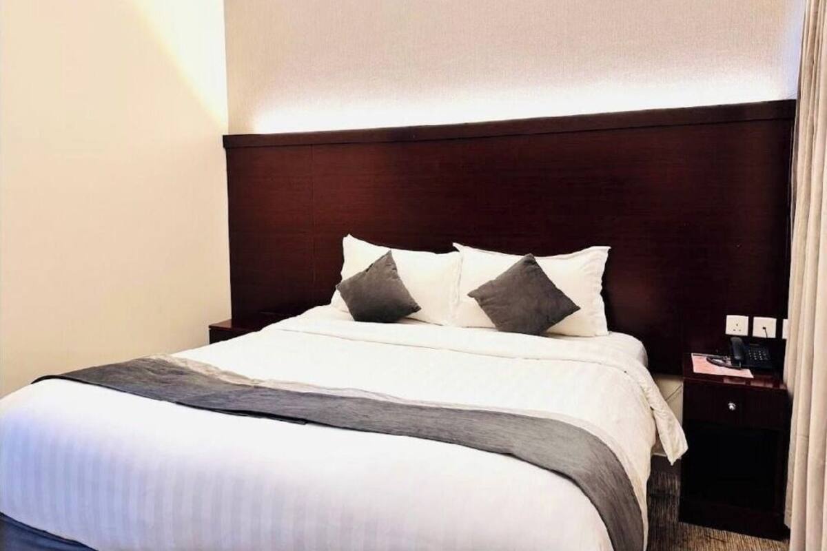 A hotel room with a large bed, a wooden headboard, two pillows, a bedside table with a phone, and a nightstand.