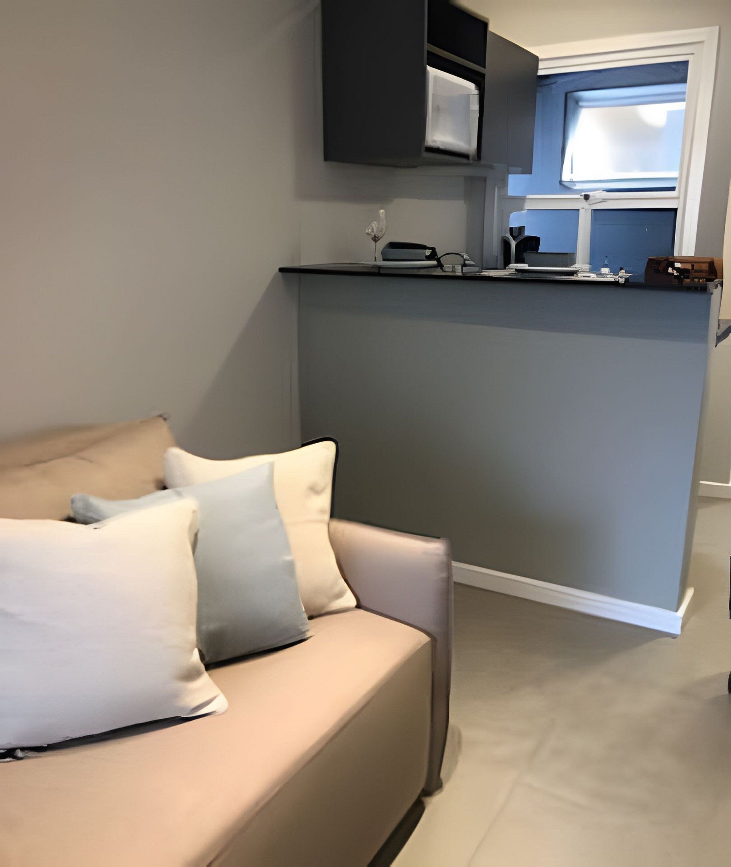 Un salon moderne avec un canapé beige, un petit coin cuisine doté d’un comptoir et d’appareils électroménagers, et une fenêtre laissant entrer la lumière naturelle.