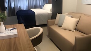 A modern hotel room with a beige sofa, a wooden side table, a blue chair, and a bed with white bedding. - Belgravia  (Porto Alegre)