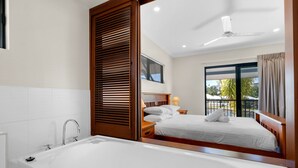 Un dormitorio moderno con una bañera blanca grande, un cabecero de madera, una cama con ropa de cama blanca, una mesita de noche con lámpara y una ventana con vistas a la vegetación.