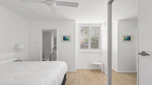 Un dormitori modern amb llit, ventilador de sostre, finestra amb persianes i una porta que condueix a una altra habitació.