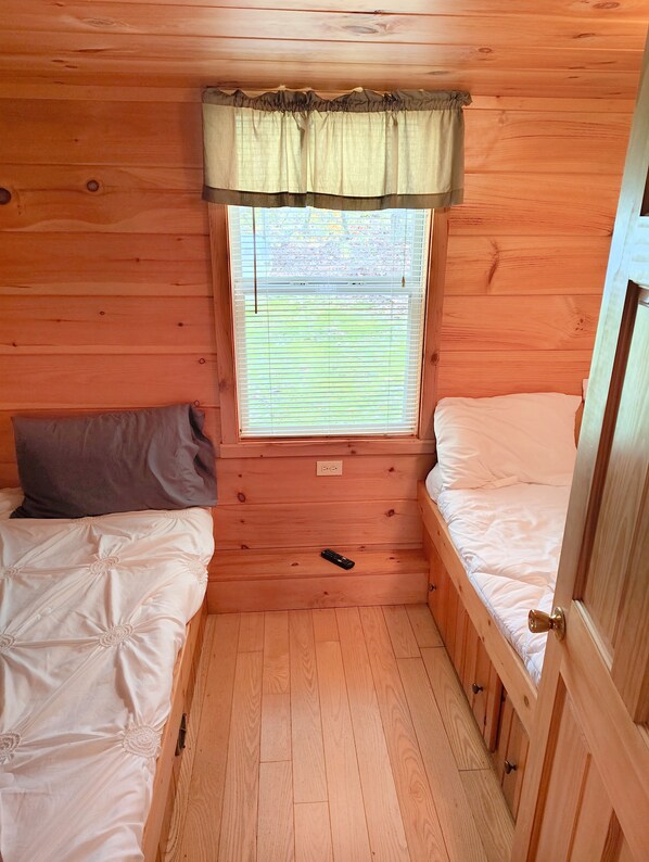 A small wooden cabin room with two beds, a window with blinds, and a door. - Pittsburgh Roaring Run RV Resort (Champion)