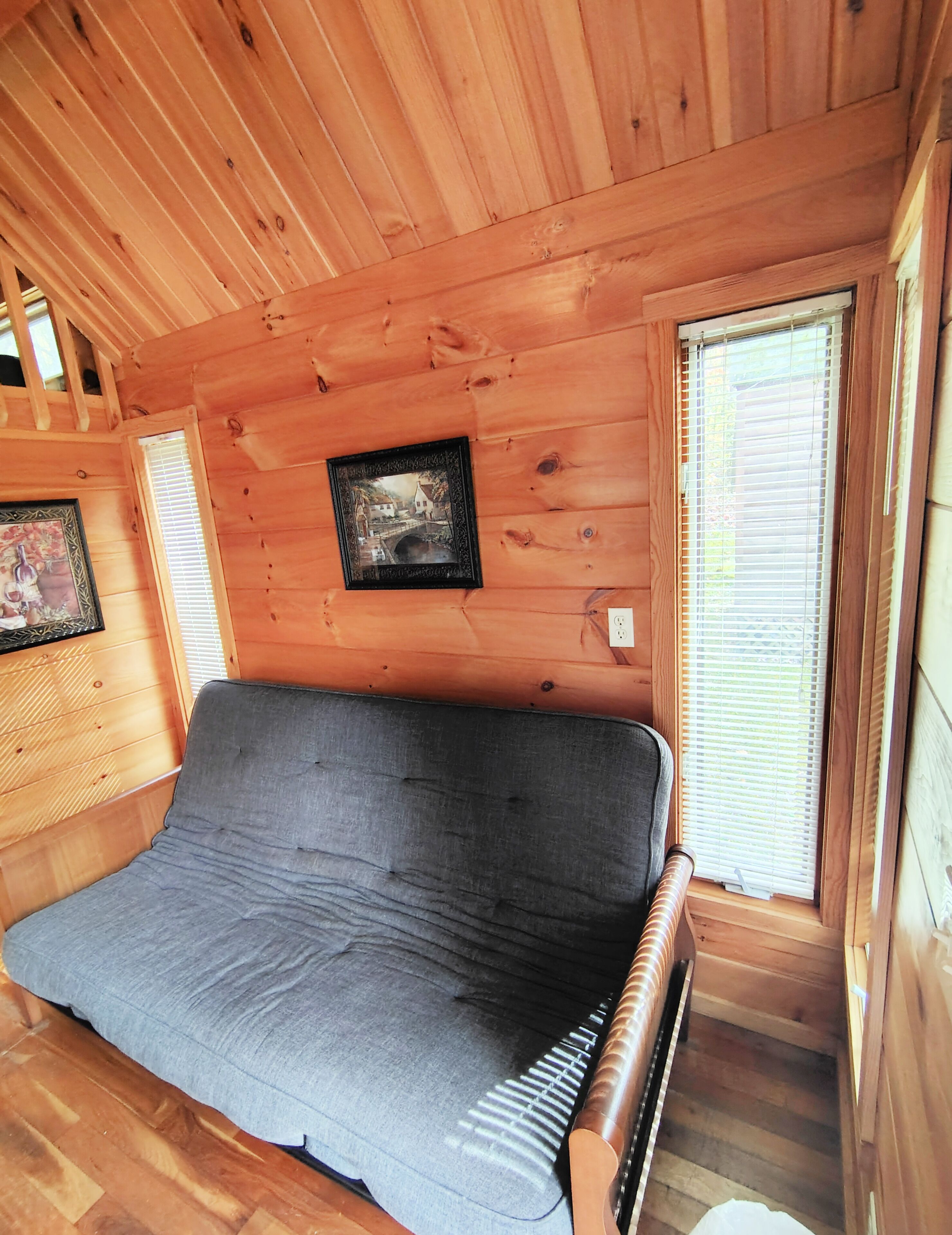 A wooden cabin interior with a sofa, wooden walls, a framed picture, and a window with blinds.