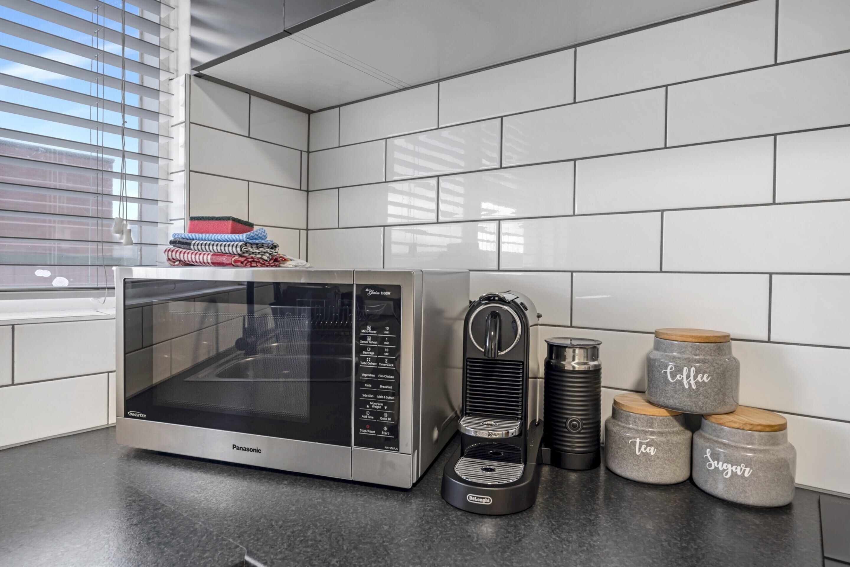A kitchen with a Panasonic microwave, a coffee maker, and three jars labeled "Coffee," "Tea," and "Sugar."