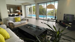 Una sala de estar moderna con sofá, mesa de centro y televisor, ventanas amplias con vista a la piscina, y una zona de cocina.