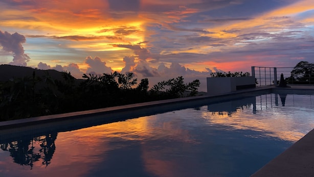 Una piscina con vista al atardecer, rodeada de follaje y una estructura de edificio blanca.