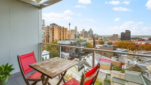 A balcony with a wooden table and red chairs, overlooking a cityscape with various buildings and a clear sky. - Cosy Studio With City Views (Sydney)