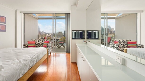 A modern bedroom with a large bed, a glass partition, a TV, and a city view. - Cosy Studio With City Views (Sydney)