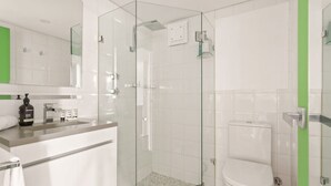 A modern bathroom with a glass-enclosed shower, a white toilet, and a vanity with a mirror and soap dispenser. - Cosy Studio With City Views (Sydney)
