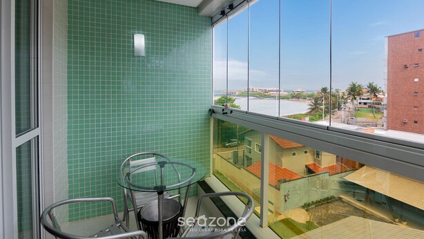 A balcony with a glass table and chairs, a green tiled wall, and a view of buildings and a body of water. - Apt w/ Balcony and sea View in Guarapari Cmd0311 (Guarapari)