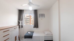 A small, clean room with a bed, a ceiling fan, a window with curtains, a coat rack, and a wall-mounted air conditioning unit. - Apt w/ Balcony and sea View in Guarapari Cmd0311 (Guarapari)