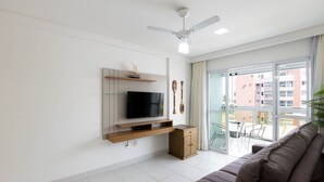 A modern living room with a flat-screen TV, wooden shelves, a ceiling fan, and a balcony with a view of other buildings. - Apt w/ Balcony and sea View in Guarapari Cmd0311 (Guarapari)