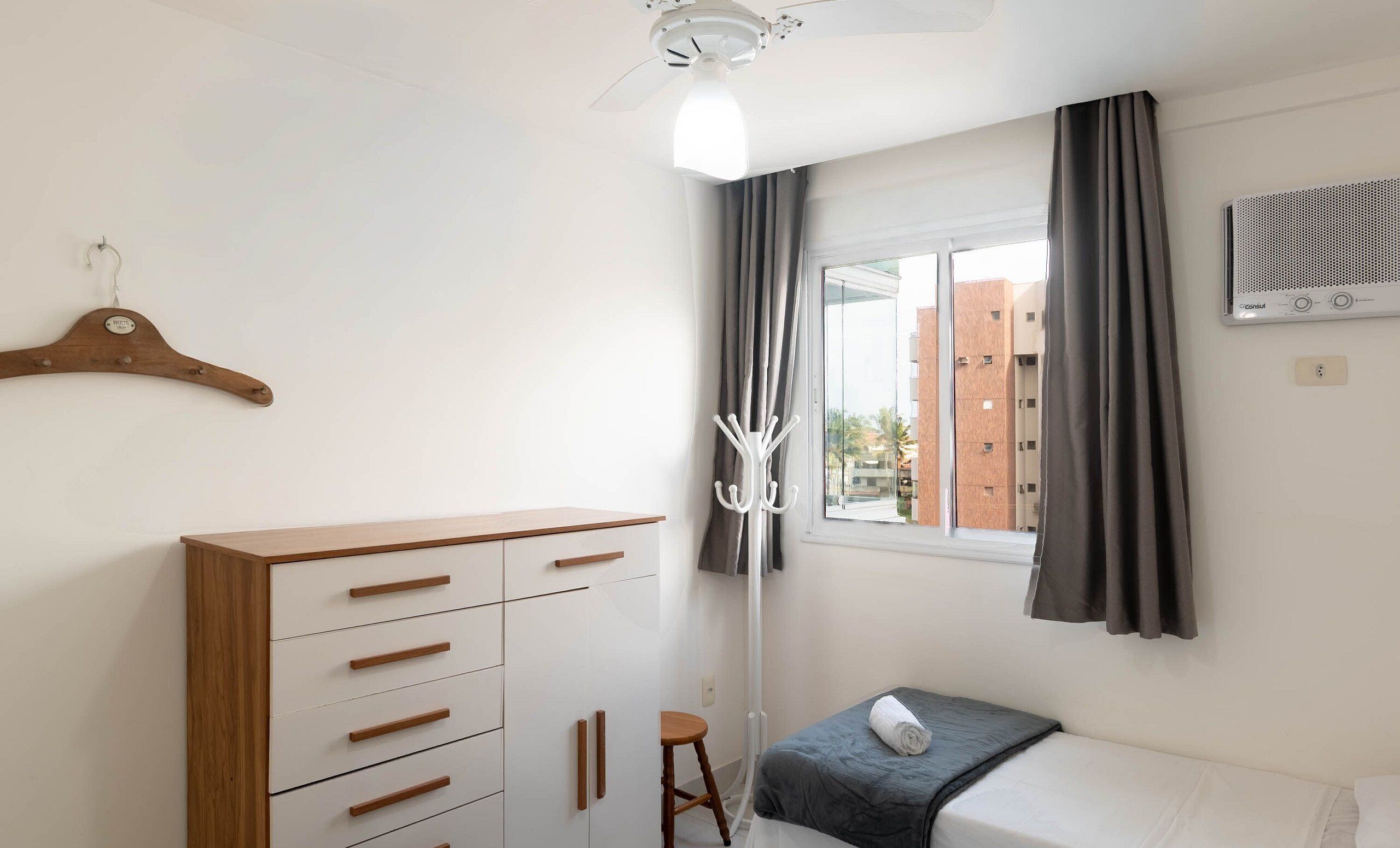 A bedroom with a wooden dresser, a white bed, a grey blanket, a white air conditioning unit, and a view of buildings outside the window.