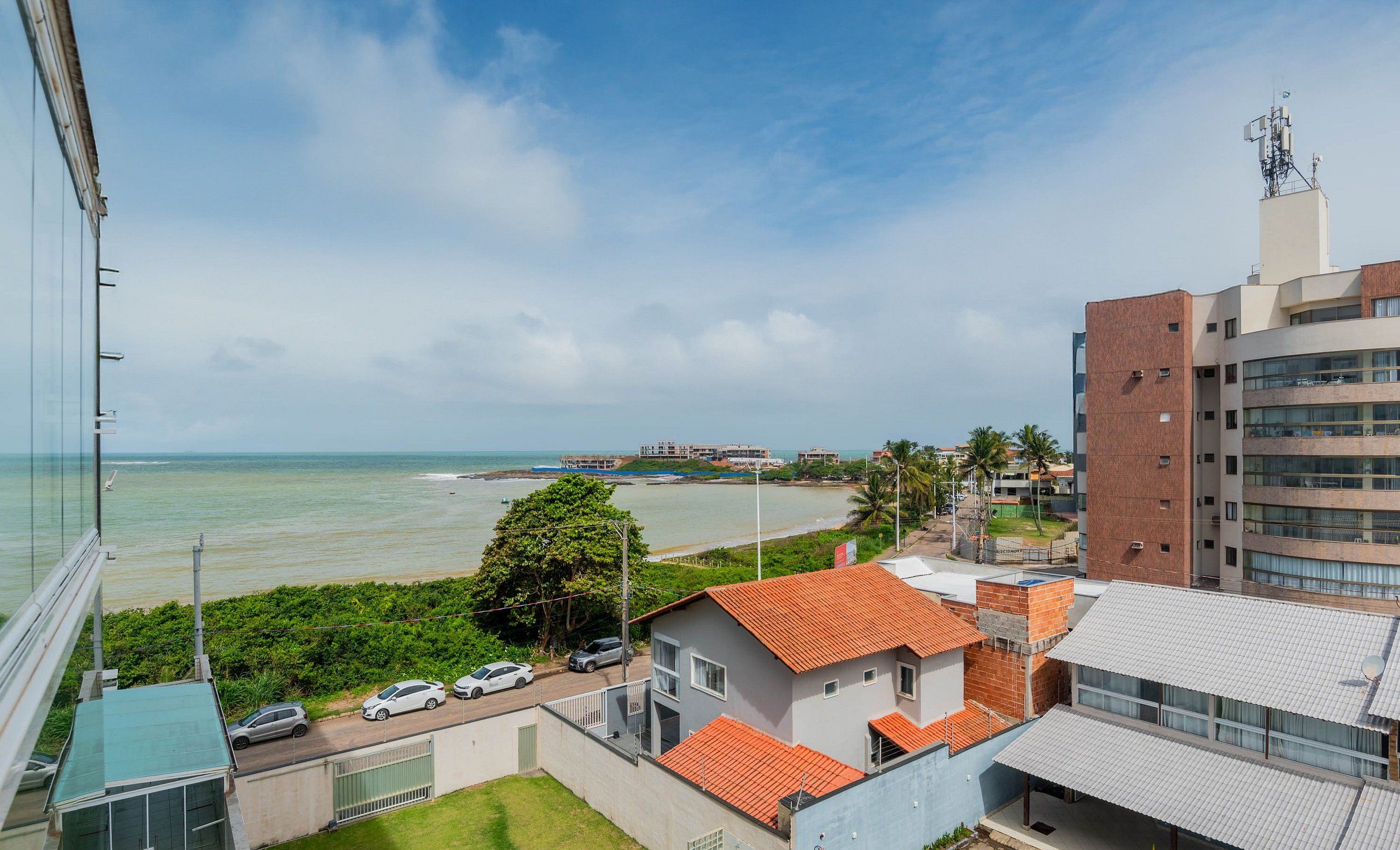 A coastal view with a building featuring a red-tiled roof, a parking area, and a clear view of the sea.