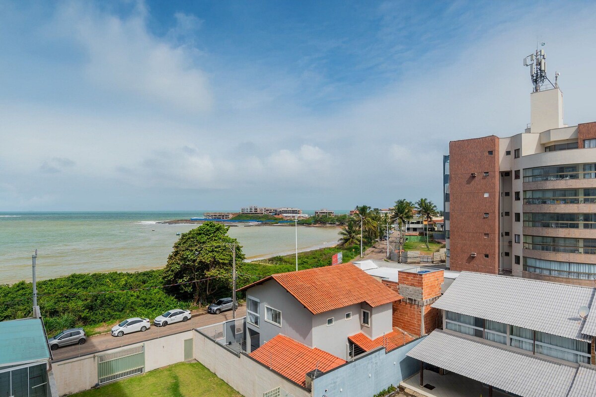 Vista costera con un edificio de techo de tejas rojas, un estacionamiento y una vista despejada del mar.