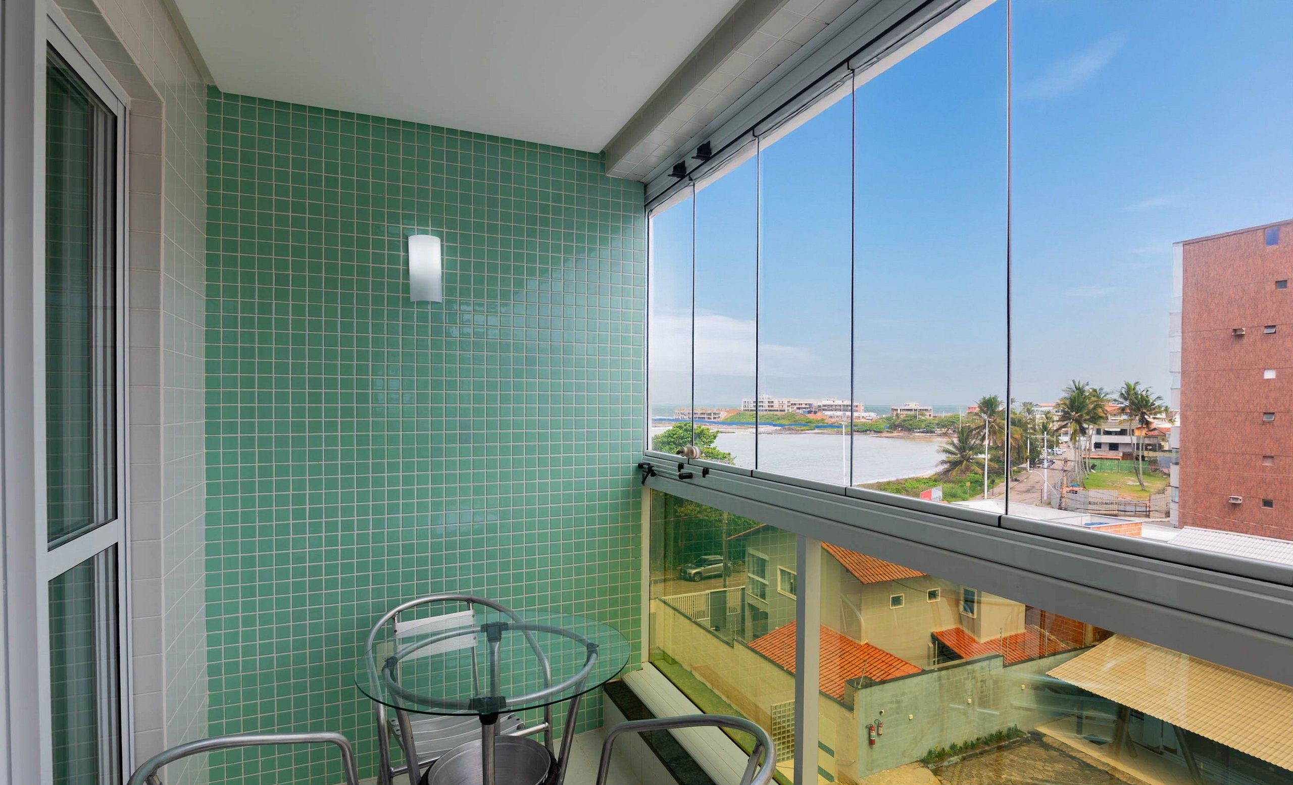 A balcony with a glass table and chairs, a green tiled wall, and a view of buildings and a body of water.