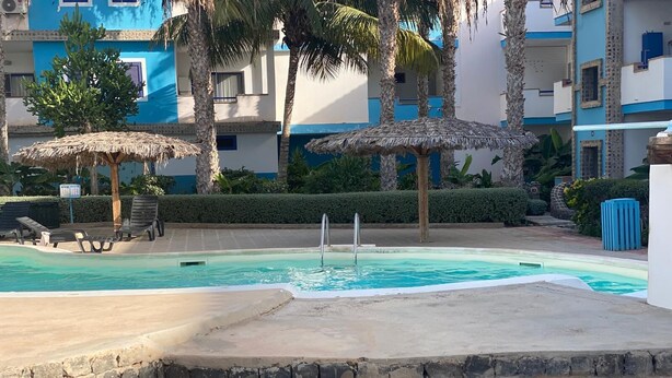 A swimming pool with lounge chairs and thatched umbrellas, surrounded by palm trees and a building with blue and white walls.
