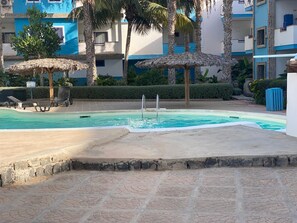 A swimming pool with lounge chairs and thatched umbrellas, surrounded by palm trees and a building with blue and white walls. - Marialva Djadsal (Santa Maria)