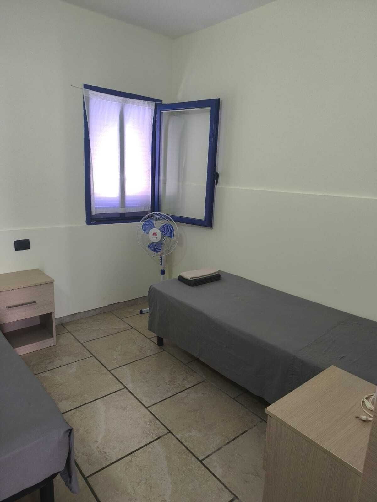 Une petite pièce, peu meublée, avec un lit simple, une table de chevet, un ventilateur sur pied et une fenêtre avec des rideaux.