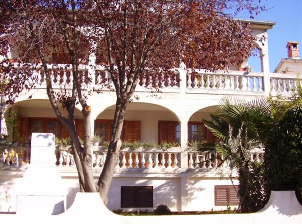 Een gebouw met twee verdiepingen, een witte gevel, meerdere balkons met sierhekwerk en een boom op de voorgrond.