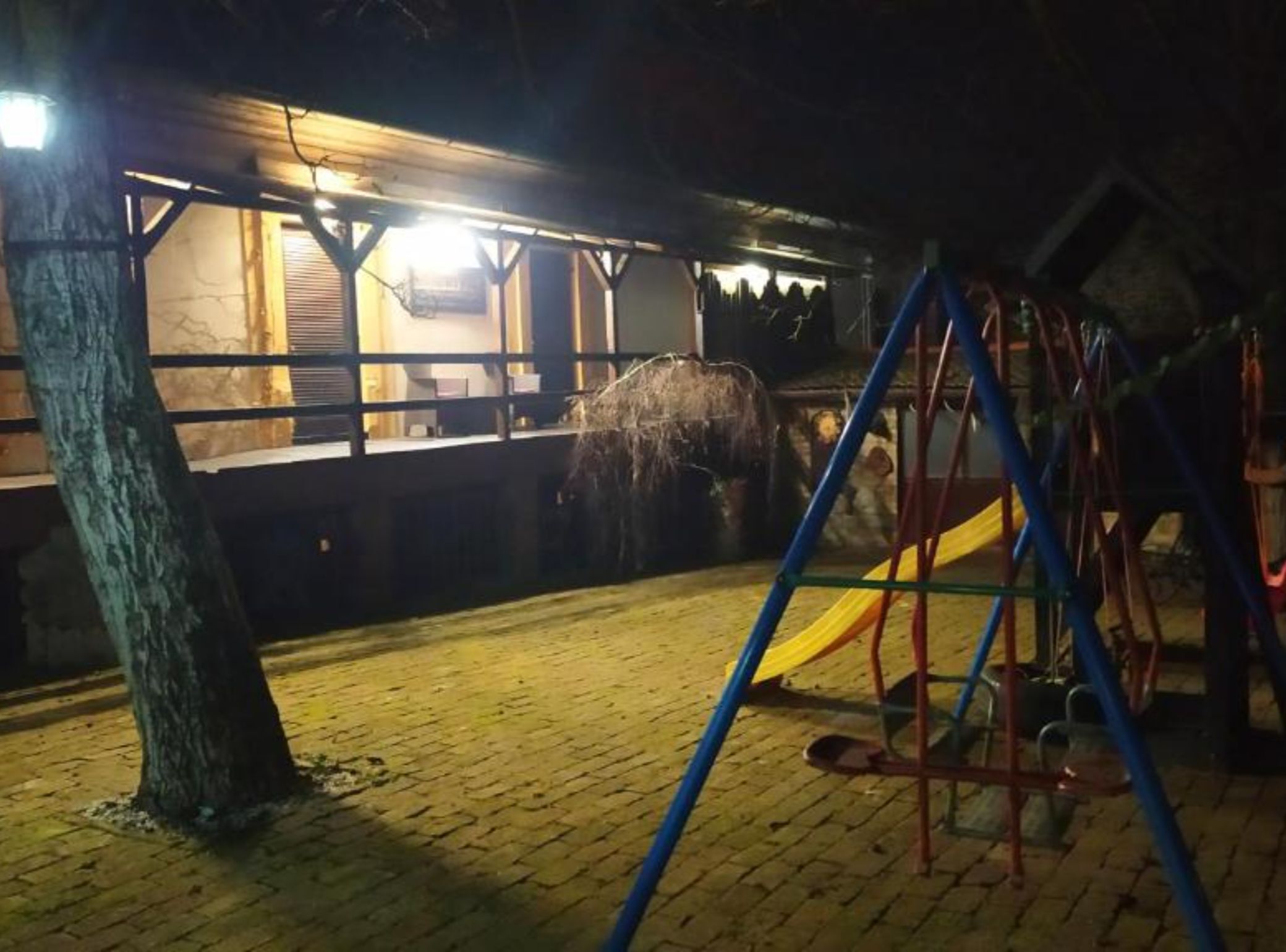 Uma área externa com piso de tijolos, uma árvore e um parquinho com um escorregador.