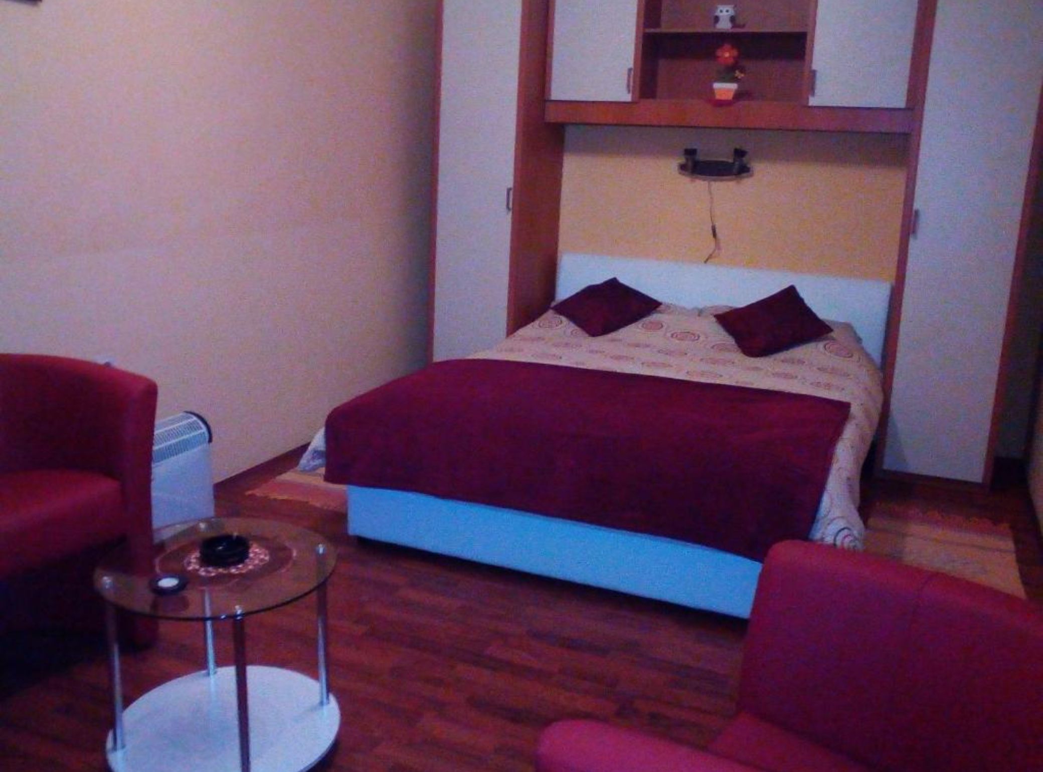 A small bedroom with a bed, two red chairs, a small table, and a heater.