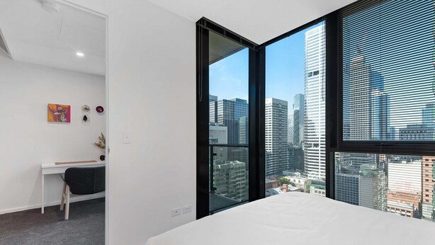 Une chambre moderne avec un grand lit, un bureau et une vue sur le paysage urbain grâce à de grandes fenêtres du sol au plafond.