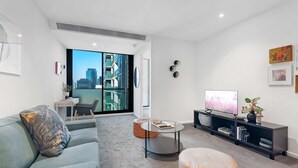 Un salon moderne avec un canapé gris, une table basse ronde et un téléviseur posé sur un meuble noir. Il y a un balcon avec vue sur la ville.