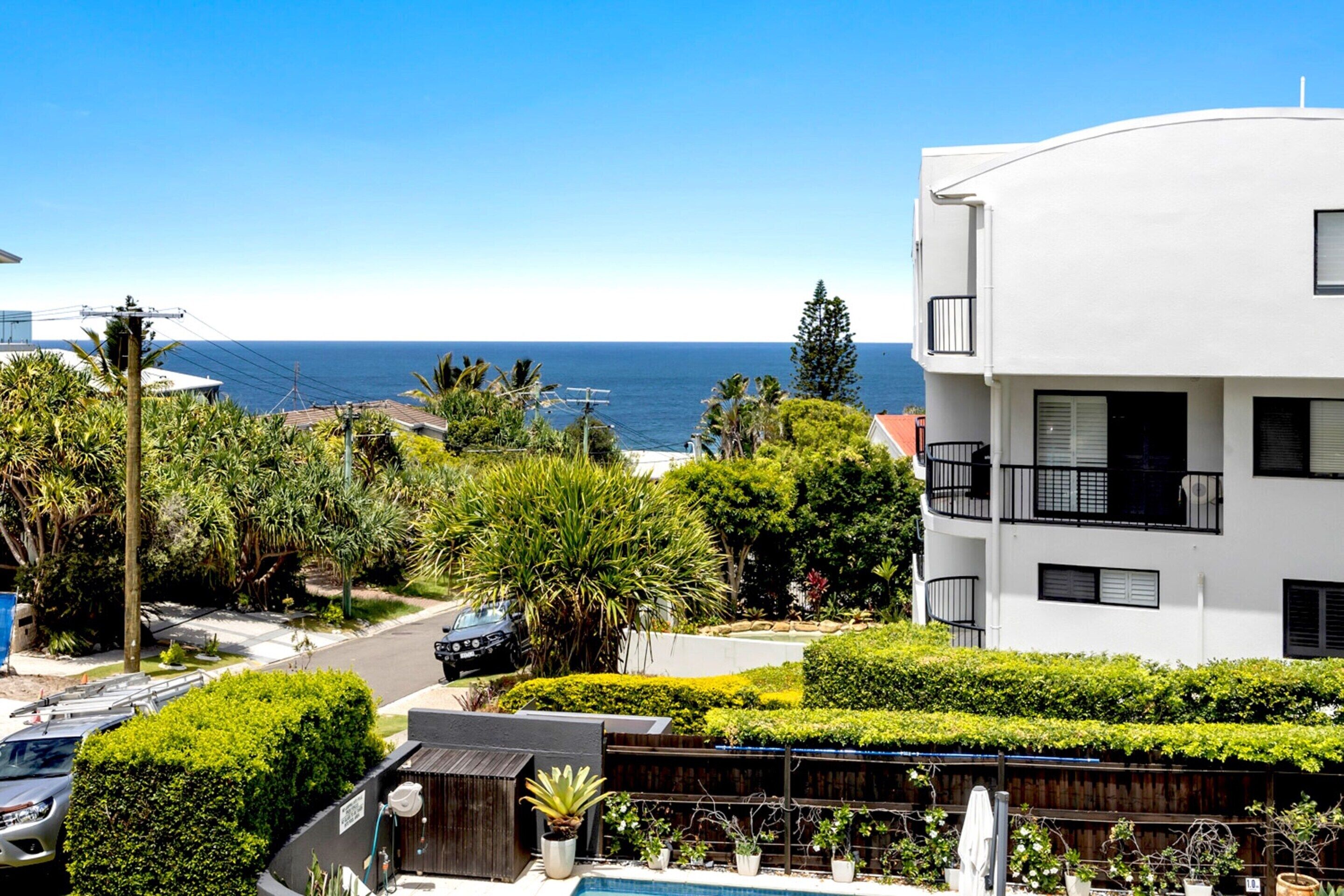 A modern white building with balconies, surrounded by lush greenery and a view of the ocean.