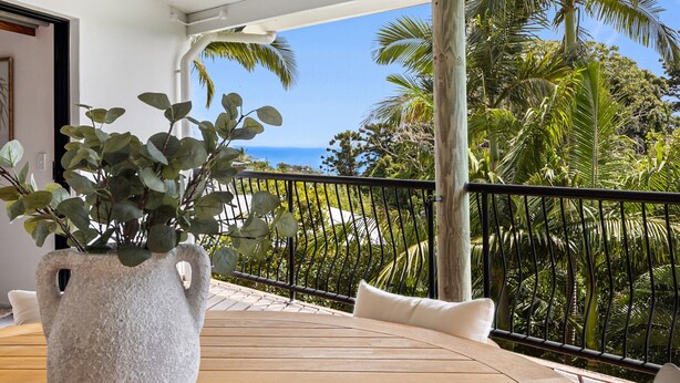 Ein Balkon mit Blick auf das Meer, eine Steinvase mit Pflanzen und ein Holztisch mit weißen Kissen.