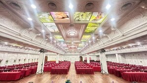 Ein großer Saal mit Reihen aus roten Tischen und Stühlen, einer verzierten Decke mit Leuchten und einer zentralen Bühne.