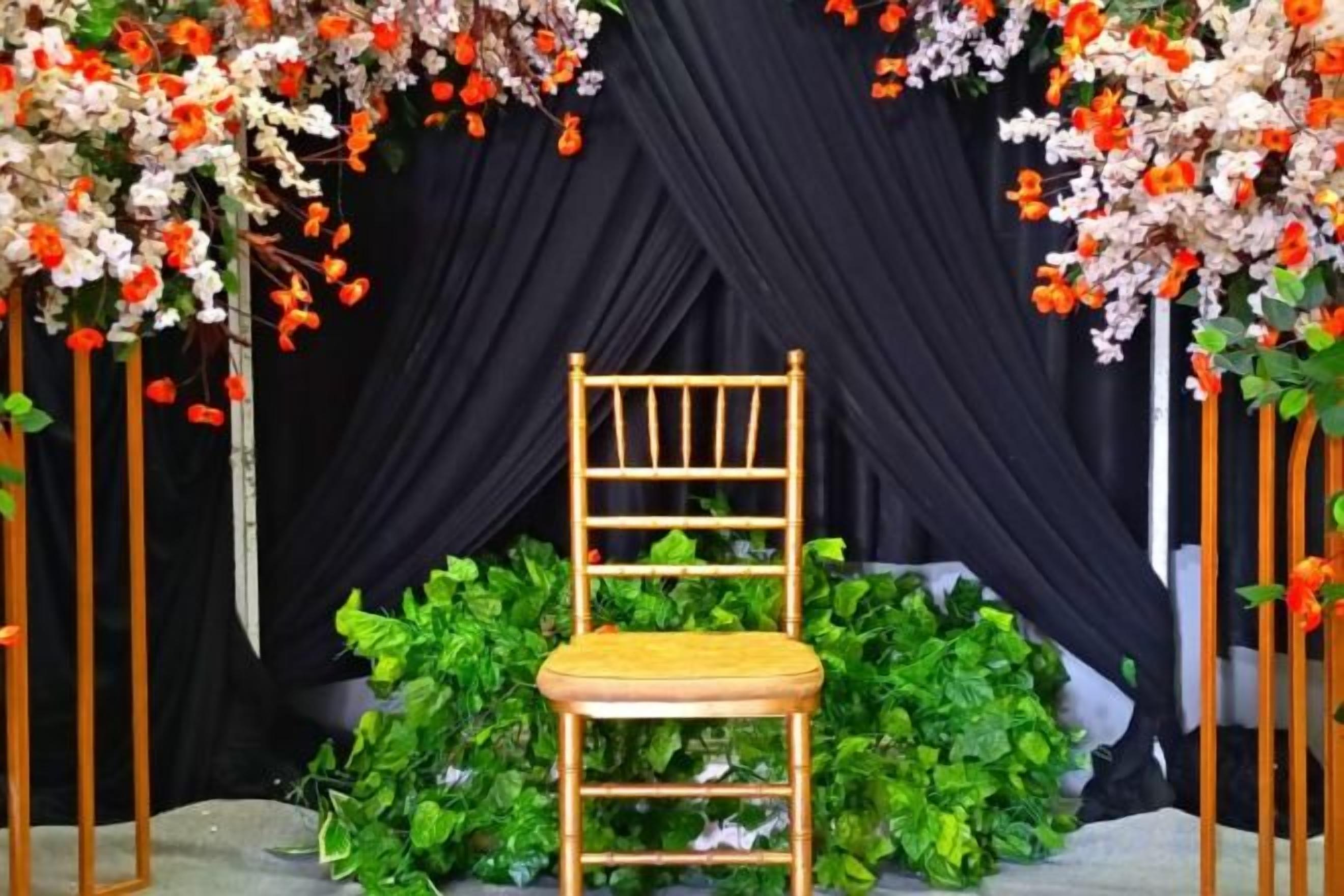 A wooden chair with a cushion in front of a black draped backdrop, surrounded by green foliage and floral arrangements.
