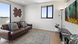 A modern living room with a brown leather sofa, a patterned rug, a large abstract painting, and a view of the city through a window. - Bright City-view Retreat in the Heart of Preston (Melbourne)