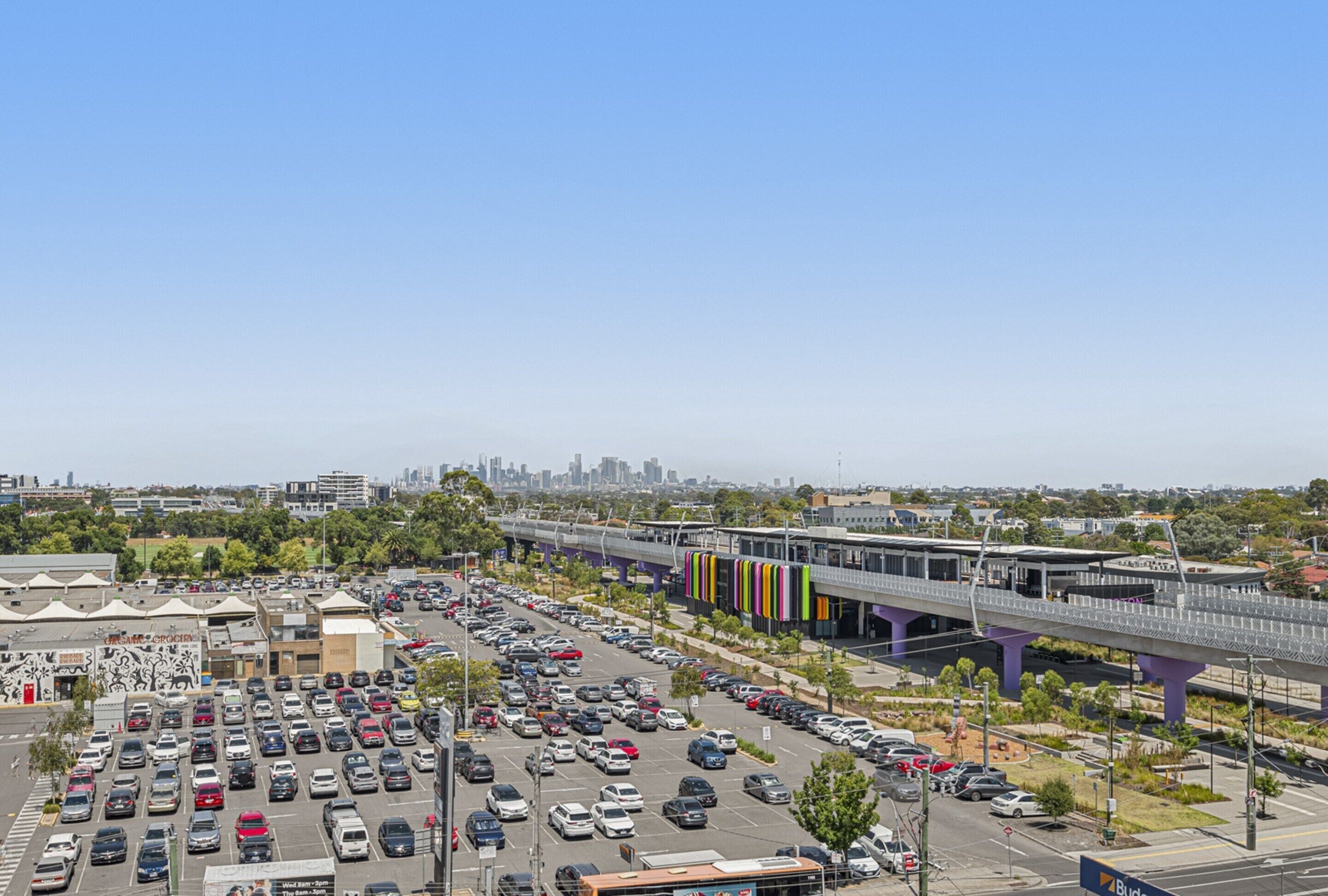 A parking lot with multiple rows of parked cars, a pedestrian overpass, and a city skyline in the background.