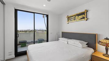 A bedroom with a large bed, a nightstand, a lamp, a sliding glass door to a balcony, and a cityscape view.
