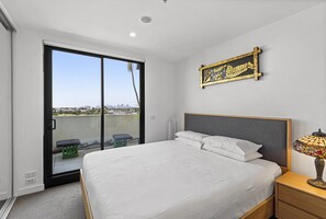 A bedroom with a large bed, a nightstand, a lamp, a sliding glass door to a balcony, and a cityscape view. - Bright City-view Retreat in the Heart of Preston (Melbourne)