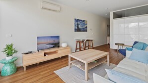 A modern living room with a wooden floor, a white sofa, a coffee table, a TV, a painting, and a small table with stools. - Stylish North Melbourne Stay (Melbourne)