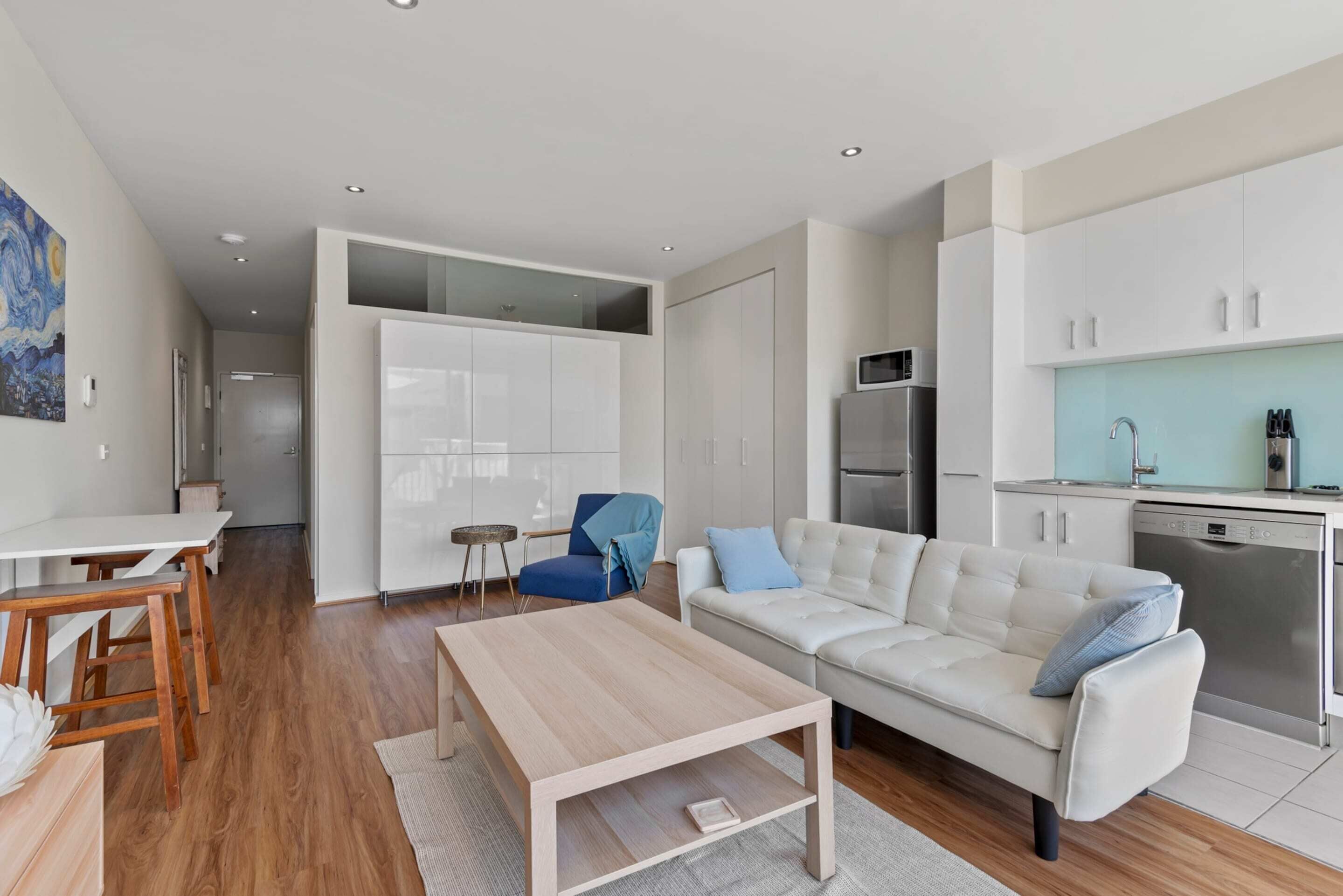 A modern living room with a sofa, coffee table, and dining area. There is a kitchen with white cabinets and a microwave.