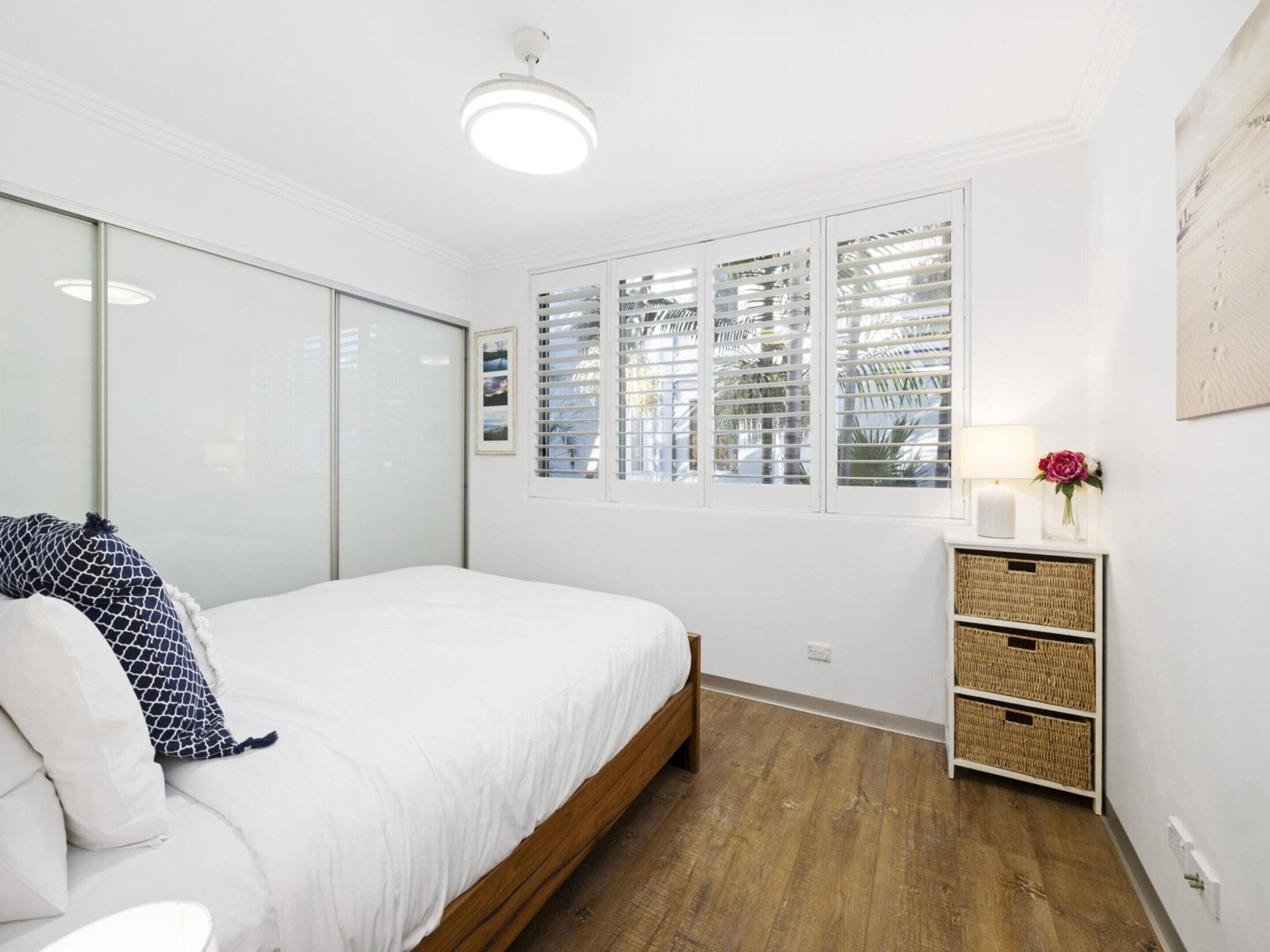 A bedroom with a bed, a bedside table with a lamp and flowers, a window with blinds, and a wall-mounted shelf with baskets.