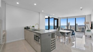 Une cuisine moderne dotée d’un îlot central, d’un coin repas avec une table et des chaises, et de grandes fenêtres offrant une vue sur la ville.