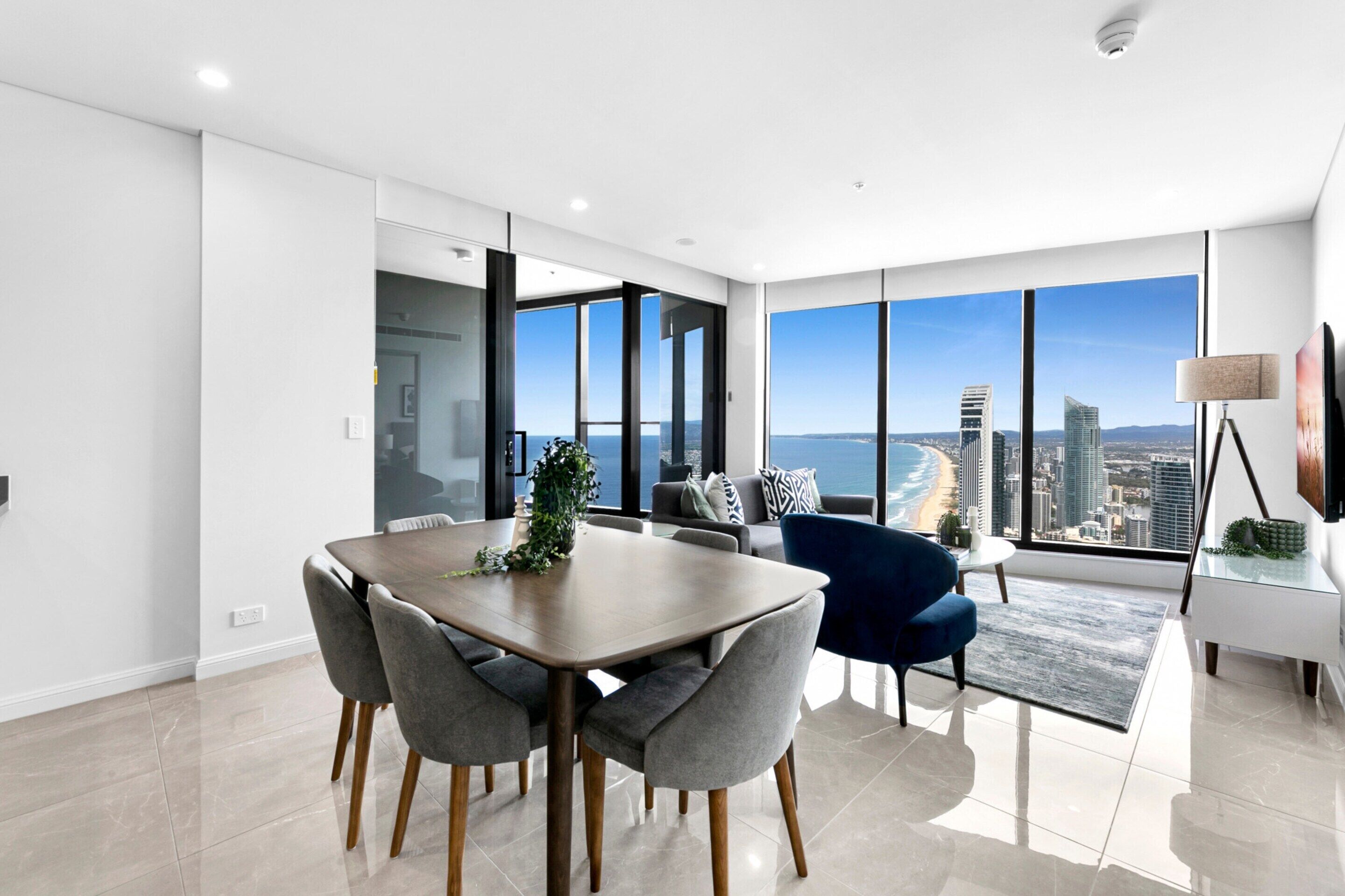 Un comedor moderno con una mesa grande y sillas, vista a la ciudad y al océano a través de amplias ventanas, y una decoración minimalista.