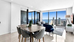 Un espace repas moderne avec une grande table et des chaises, une vue sur la ville et l’océan grâce à de larges fenêtres, et un décor minimaliste.