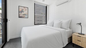 A modern bedroom with a bed, a nightstand, a desk lamp, a window with blinds, and a framed picture on the wall. - Stylish West End Stay With Rooftop Pool & BBQ (Brisbane)