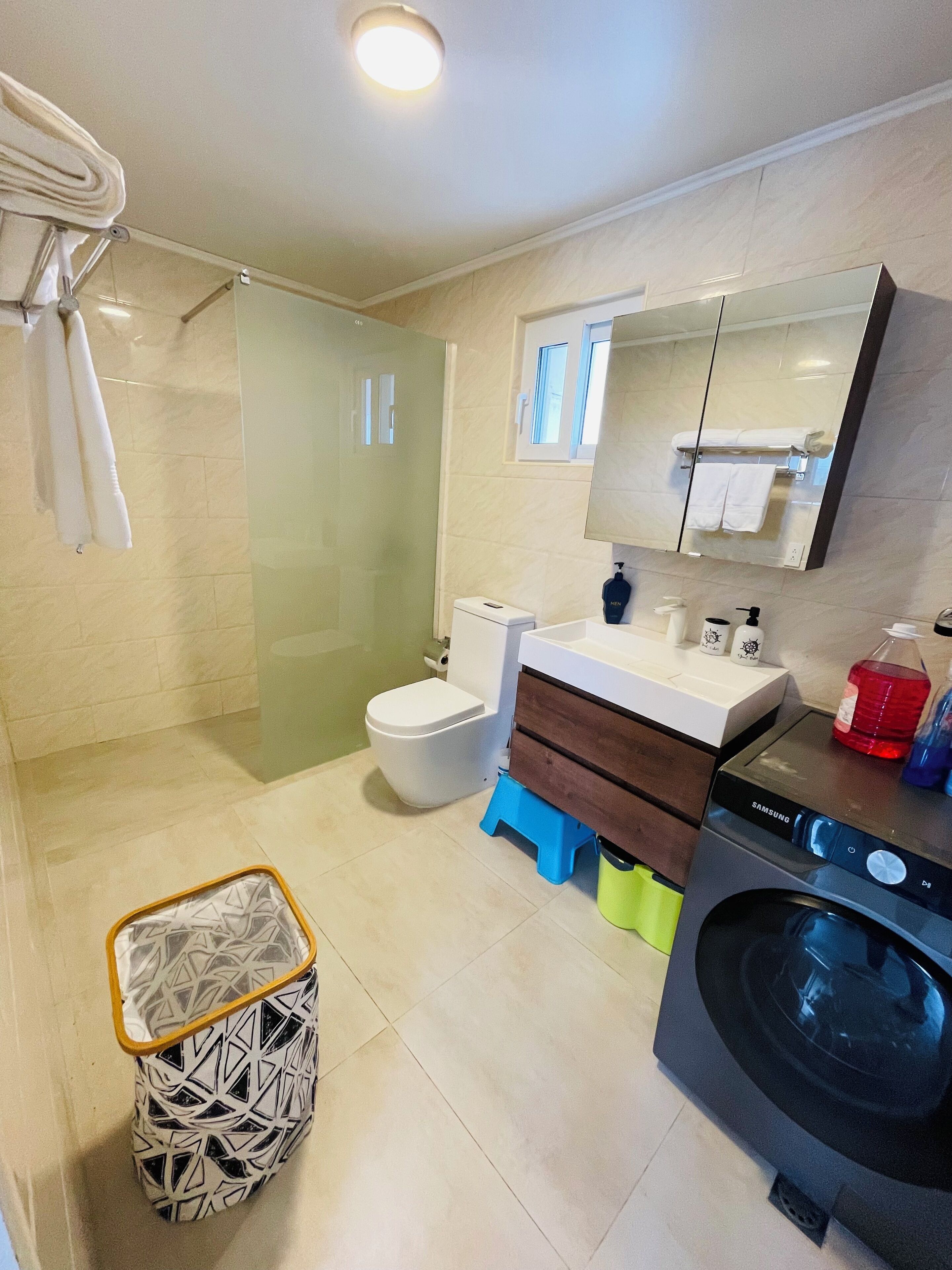 A modern bathroom with a glass shower enclosure, a toilet, a sink with a mirror, a washing machine, and a patterned trash bin.