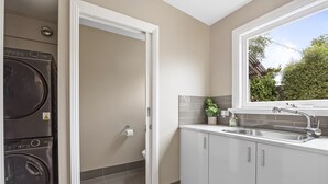 A laundry room with stacked washers and dryers, a white door, and a kitchen area with a sink and window. - Lovely 3-bed With Alfresco Dining in Prime Suburb (Ballarat)