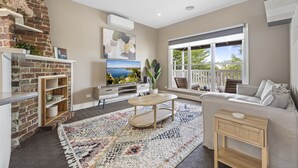 A modern living room with a brick fireplace, a large window with a view, a sofa, and a coffee table. - Lovely 3-bed With Alfresco Dining in Prime Suburb (Ballarat)