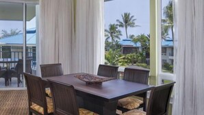 Un coin repas avec une table et des chaises, de grandes fenêtres avec des rideaux et une vue sur des palmiers et des immeubles.