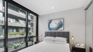 Une chambre moderne avec un grand lit, deux tables de chevet avec des lampes, un tableau abstrait accroché au mur, et une vue sur un immeuble doté de balcons et de plantes, visible à travers de grandes fenêtres du sol au plafond.