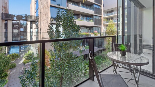 Un balcon doté d’une balustrade en verre, une petite table avec un pot de fleurs, et une vue sur un paysage urbain avec des bâtiments et de la verdure.