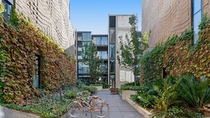Une ruelle étroite, avec un vélo garé sur le côté gauche, bordée de bâtiments en briques dont les murs sont recouverts de lierre, et un jardinier avec différentes plantes sur le côté droit.